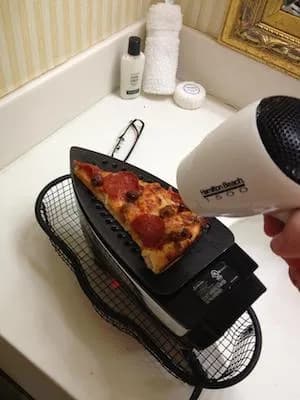 A humorous photo of a pizza slice being heated with a clothes iron and hair dryer, illustrating a hacky improvised solution.